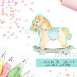 May include: A 3D printed cookie cutter shaped like a rocking horse. The horse is light brown with a blue saddle and a light brown mane. The cookie cutter is on a white background with colorful sprinkles.