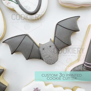 Puede incluir: Un cortador de galletas en forma de murciélago gris con las alas extendidas, rodeado de otras galletas decoradas. El texto "CUSTOM 3D PRINTED COOKIE CUTTER" es visible. El cortador de galletas es probablemente para Halloween o repostería temática.