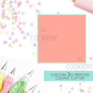 May include: A coral pink 3D printed cookie cutter with the text "The Cookiery Made by Mama" in white. The cookie cutter is surrounded by pastel pink, yellow, and blue heart sprinkles and piping bags.