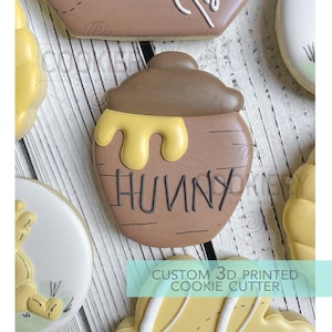 May include: A brown cookie shaped like a honey pot with the word "HUNNY" written on it in black. Yellow icing drips from the top of the pot. The image also shows other cookies and the text "CUSTOM 3D PRINTED COOKIE CUTTER".