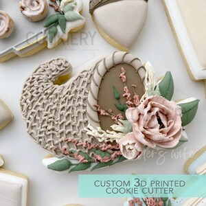 May include: A 3D printed cookie cutter shaped like a cornucopia with a floral design. The cornucopia is brown and white, and the flowers are pink, white, and green. The text "CUSTOM 3D PRINTED COOKIE CUTTER" is printed below the cookie cutter.
