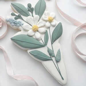 Op de afbeelding: Een witte koek versierd met een boeket bloemen. De bloemen zijn wit met gele centra, omringd door groene bladeren en stelen. De koek heeft de vorm van een boeket en is versierd met lichtblauwe accenten.
