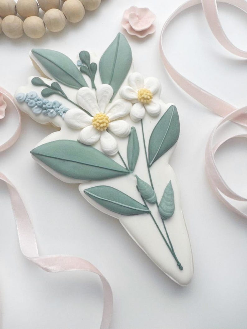 Op de afbeelding: Een witte koek versierd met een boeket bloemen. De bloemen zijn wit met gele centra, omringd door groene bladeren en stelen. De koek heeft de vorm van een boeket en is versierd met lichtblauwe accenten.