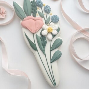 Op de afbeelding: Een witte, bloemvormige koek versierd met een boeket pastelkleurige bloemen en bladeren. Het boeket bevat een roze tulp, een witte madelief en blauwe pomponbloemen, met groene bladeren. Roze lint en houten kralen op de achtergrond.