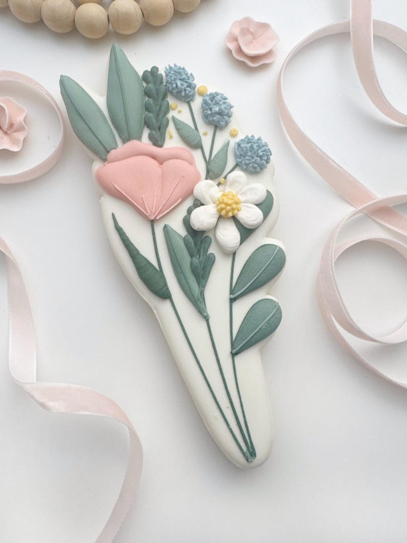 Op de afbeelding: Een witte, bloemvormige koek versierd met een boeket pastelkleurige bloemen en bladeren. Het boeket bevat een roze tulp, een witte madelief en blauwe pomponbloemen, met groene bladeren. Roze lint en houten kralen op de achtergrond.