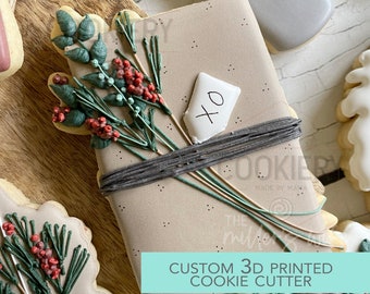 May include: A 3D printed cookie cutter in the shape of a wrapped gift box with a sprig of greenery and a small tag that says "xo". The cookie cutter is made of white plastic and is designed for making cookies.