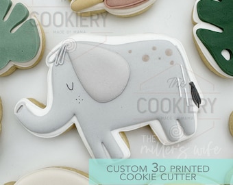 May include: A gray 3D printed cookie cutter shaped like an elephant. The cutter has a small tail, a trunk, and a small U-shaped mark on the side. The text "The Cookiery Made by Mama" is visible in the background.