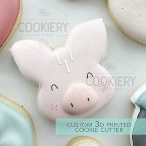 Könnte beinhalten: Ein rosa Schweinchen-Ausstecher mit einer weißen Schnauze und Augen. Der Ausstecher ist 3D-gedruckt und trägt den Text "The Cookiery Made by Mama" oben und unten.