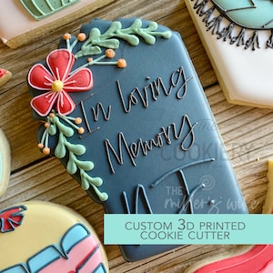 Pode incluir: Um cortador de biscoitos impresso em 3D de cor preta com a forma de uma lápide com as palavras "In loving memory" escritas em glacê branco. A lápide é decorada com uma flor vermelha e folhas verdes.