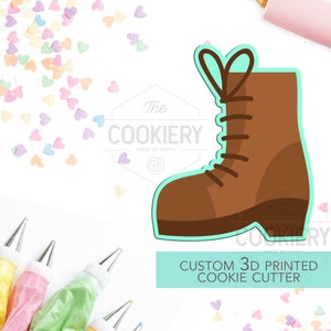 May include: A brown boot cookie cutter with a light blue outline. The cutter is 3D printed and is a custom design. The text "The Cookiery Made by Mama" is in the background.