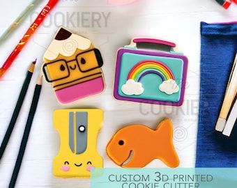 May include: Four 3D printed cookie cutters in the shape of a pencil, a lunchbox with a rainbow, a pencil sharpener, and a fish. The cutters are on a white surface with pencils and a blue napkin.