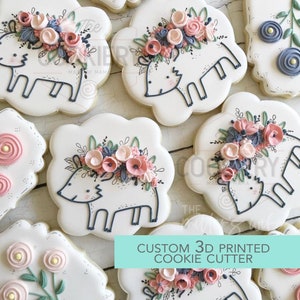 Op de afbeelding: Close-up van versierde suikerkoekjes. Elk koekje is wit en heeft een hertenontwerp met blauwe contouren, versierd met bloemaccenten in roze, blauw en groen. De koekjes zijn gerangschikt op een lichtgekleurd oppervlak. De tekst op de afbeelding luidt "CUSTOM 3D PRINTED COOKIE CUTTER."