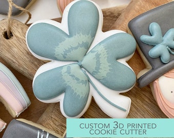 Könnte beinhalten: Ein 3D-gedruckter Ausstecher in Form eines vierblättrigen Kleeblatts. Der Ausstecher ist weiß mit einem hellblauen Kleeblatt-Design. Der Text "CUSTOM 3D PRINTED COOKIE CUTTER" ist am unteren Rand des Bildes sichtbar.