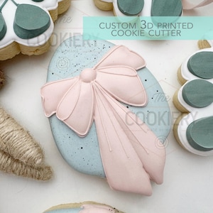May include: A decorated sugar cookie in the shape of an egg with a pink bow. The cookie has a light blue background with black speckles. The text "CUSTOM 3D PRINTED COOKIE CUTTER" is in a light blue rectangle.