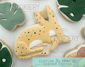 May include: A 3D printed cookie cutter in the shape of a cheetah, with a light brown body, black spots, and a white chest. The text "CUSTOM 3D PRINTED COOKIE CUTTER" is printed on a light blue background.