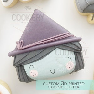 Puede incluir: Un cortador de galletas impreso en 3D con forma de bruja con un sombrero morado, cabello gris y una cara azul claro. El cortador de galletas está decorado con detalles rosas y negros. El texto "The Cookery Made by Mama" es visible en el cortador de galletas.