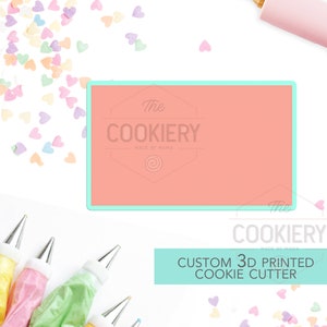 May include: A light pink cookie cutter with the text "The Cookiery Made by Mama" in a white circle. The cookie cutter is on a white background with colorful heart sprinkles and piping bags.
