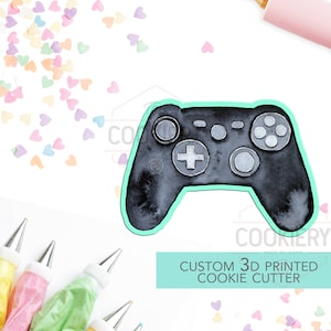 Może przedstawiać: Czarno-szara 3D drukarka do ciasteczek w kształcie kontrolera do gier wideo. Wycinarka znajduje się na białym tle z kolorowymi posypkami i woreczkami do dekorowania.
