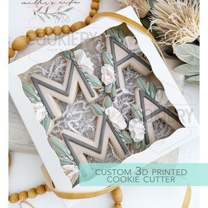 May include: A white box with a gold ribbon contains two 3D printed cookie cutters shaped like the letters "M" and "A". The cutters are decorated with floral details and are labeled "CUSTOM 3D PRINTED COOKIE CUTTER".
