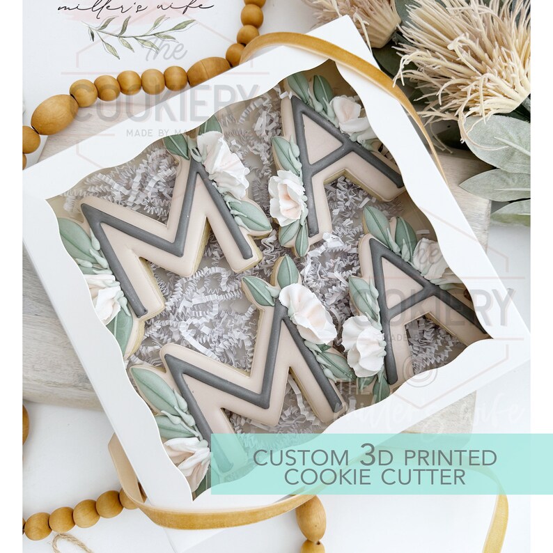 May include: A white box with a gold ribbon contains two 3D printed cookie cutters shaped like the letters "M" and "A". The cutters are decorated with floral details and are labeled "CUSTOM 3D PRINTED COOKIE CUTTER".
