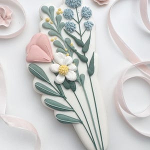 Op de afbeelding: Een versierd suikerkoekje in de vorm van een boeket. De koek heeft een roze tulp, witte en blauwe bloemen en groene bladeren. Roze lint en een kleine roze bloem staan op de achtergrond.