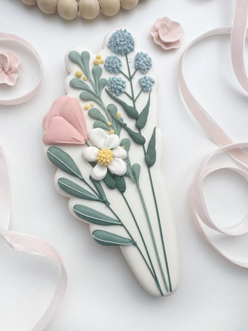 Op de afbeelding: Een versierd suikerkoekje in de vorm van een boeket. De koek heeft een roze tulp, witte en blauwe bloemen en groene bladeren. Roze lint en een kleine roze bloem staan op de achtergrond.