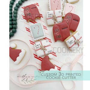 May include: A collection of decorated sugar cookies shaped like red mailboxes with HO HO HO text, white envelopes addressed to the North Pole with gingerbread men, candy canes, and red Santa hats, arranged with green beads and ribbons.