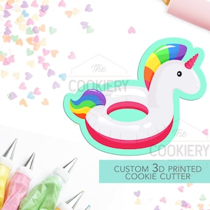 Puede incluir: Un cortador de galletas impreso en 3D con forma de flotador de unicornio. El flotador es blanco con una crin y cola de arcoíris. El cortador de galletas está sobre un fondo blanco con chispas y bolsas de decoración.