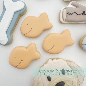May include: Overhead shot of decorated cookies in the shape of a fish, a bone, and a dog's face. The fish cookies are light orange with a smiling face. The bone cookie is white and light blue. The dog cookie has a light brown face. Text on the image reads "CUSTOM 3D PRINTED COOKIE CUTTER."
