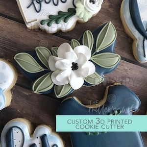 May include: A blue and green cookie cutter in the shape of a onesie with a white flower and green leaves. The text "Custom 3D Printed Cookie Cutter" is on a teal background.