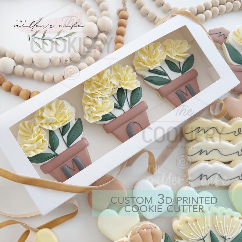 Peut inclure: Trois biscuits en forme de fleurs jaunes dans une bo&icirc;te blanche avec une fen&ecirc;tre transparente. Les biscuits sont d&eacute;cor&eacute;s de feuilles vertes et de pots bruns. Le mot "MAMAN" est &eacute;crit sur les pots. La bo&icirc;te est &eacute;tiquet&eacute;e "The Cookiery Made by Mama".
