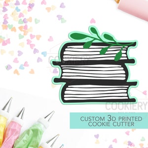 May include: A 3D printed cookie cutter in the shape of a stack of books with a green leafy branch on top. The cutter is made of white plastic and has a light blue outline. The text "CUSTOM 3D PRINTED COOKIE CUTTER" is written in white on a light blue background.