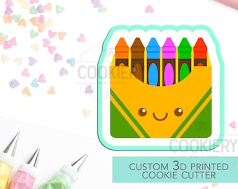 May include: A 3D printed cookie cutter shaped like a box of crayons. The box is yellow with a green lid and a smiling face. The crayons are red, orange, yellow, brown, blue, pink, and green. The cookie cutter is made by The Cookiery.
