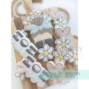 May include: A set of cookie cutters featuring a woman with a messy bun, flowers, and the word "Mom". The cutters are made of white plastic and are designed for use with cookie dough. The text "CUSTOM 3D PRINTED COOKIE CUTTER" is also visible in the image.