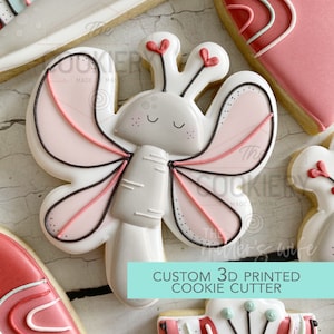 May include: A 3D printed cookie cutter in the shape of a butterfly with pink wings, a gray body, and red antennae. The text "CUSTOM 3D PRINTED COOKIE CUTTER" is printed on a turquoise background.