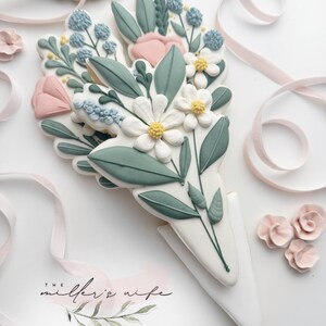 Op de afbeelding: Een versierd suikerkoekje in de vorm van een boeket bloemen. Het koekje heeft witte, roze en blauwe bloemen met groene bladeren. De basis is wit en kegelvormig. De tekst "the miller's wife" is zichtbaar.