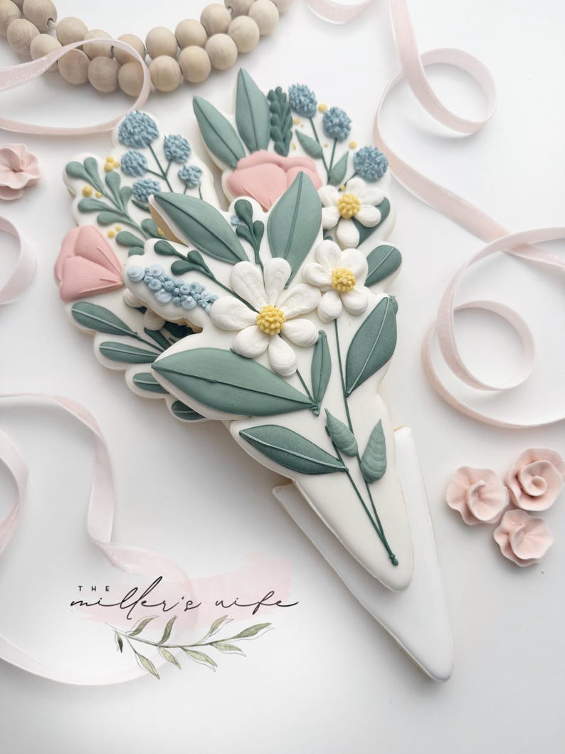 Op de afbeelding: Een versierd suikerkoekje in de vorm van een boeket bloemen. Het koekje heeft witte, roze en blauwe bloemen met groene bladeren. De basis is wit en kegelvormig. De tekst "the miller's wife" is zichtbaar.