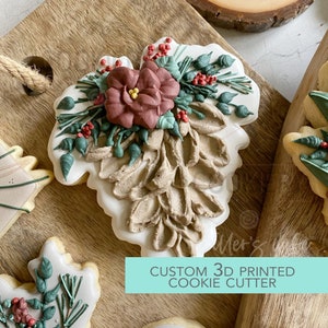 Può includere: Un tagliapasta stampato in 3D a forma di pigna con un fiore rosso e foglie verdi. Il testo "CUSTOM 3D PRINTED COOKIE CUTTER" è su uno sfondo turchese.