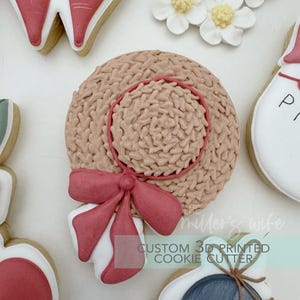 May include: A beige cookie shaped like a straw hat with a red ribbon and trim. The hat has a textured surface and is surrounded by other decorated cookies and white flowers. The text "CUSTOM 3D PRINTED COOKIE CUTTER" is visible.