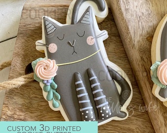 Può includere: Un tagliapasta a forma di gatto stampato in 3D di colore grigio, che tiene una rosa rosa. Il gatto ha la pancia bianca, strisce nere sulle gambe e pois bianchi sui piedi. Il testo "CUSTOM 3D PRINTED COOKIE CUTTER" è stampato su uno sfondo turchese sotto il tagliapasta.