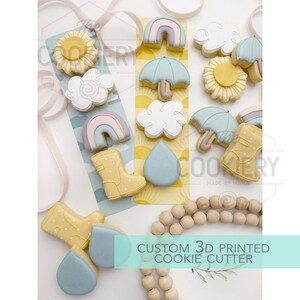 May include: A collection of custom 3D printed cookie cutters shaped like a sun, cloud, rainbow, umbrella, wellington boot, and raindrop. The cookies are decorated in pastel colours and arranged on a decorative background.