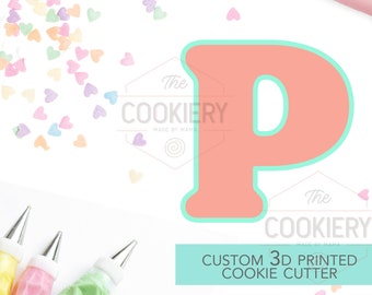 May include: A light pink 3D printed cookie cutter in the shape of the letter P. The cutter is surrounded by pastel-colored heart sprinkles and piping bags. The text "The Cookiery" is visible in the image.