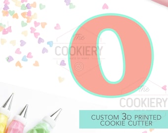 May include: A pink and green 3D printed cookie cutter in the shape of the letter O. The cutter is surrounded by pastel-colored heart sprinkles and a rolling pin. The text "The Cookiery Made by Mama" is visible in the image.