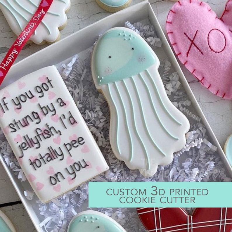 May include: A custom 3D printed cookie cutter in the shape of a jellyfish. The cutter is light blue and white with a light blue bell and white tentacles. The cutter is in a white box with white shredded paper. The box also contains a cookie decorated with pink hearts and the text "If you got stung by a jellyfish, I'd totally pee on you".