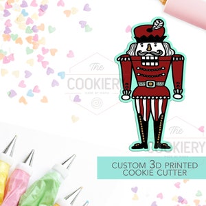 May include: A 3D printed cookie cutter in the shape of a nutcracker. The nutcracker is red, white, and black with a gold belt and boots. The cookie cutter is on a white background with colorful sprinkles and piping bags.