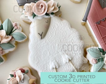 May include: A selection of decorated biscuits, including a sheep with a floral crown, a barn, and floral accents. The sheep biscuit is white with a light brown face and a flower crown. The text "CUSTOM 3D PRINTED COOKIE CUTTER" is on a teal banner.