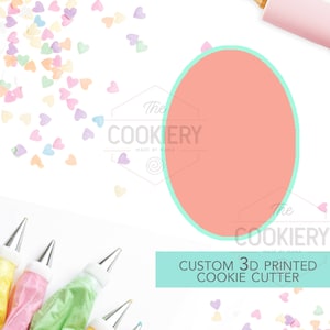 May include: A custom 3D printed cookie cutter in the shape of a heart. The cutter is pink and has a light blue outline. The image also shows a rolling pin, piping bags, and sprinkles in the shape of hearts.