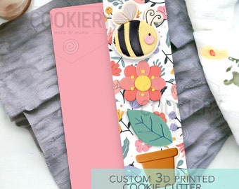 May include: A 3D printed cookie cutter featuring a bee, flower, and potted plant. The cutter is white with pink, yellow, and green accents. The text "CUSTOM 3D PRINTED COOKIE CUTTER" is printed on the bottom of the cutter.