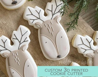 May include: Four decorated sugar cookies shaped like a reindeer head with antlers. The cookies are on a wooden surface. The text "CUSTOM 3D PRINTED COOKIE CUTTER" is on a turquoise background.