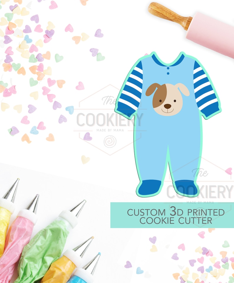 May include: Blue 3D printed cookie cutter shaped like a baby onesie with a dog face on the front. The cookie cutter is on a white background with colorful sprinkles.